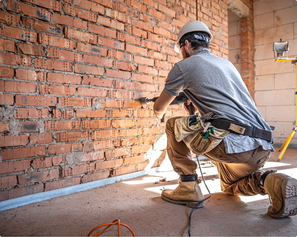 Trabajador realizando perforaciones en una pared de ladrillos expuestos, utilizando una herramienta eléctrica. Está agachado y lleva un casco de seguridad, gafas de protección y un cinturón de herramientas con diversos utensilios. El entorno muestra una obra en curso con materiales y equipos visibles en el fondo.