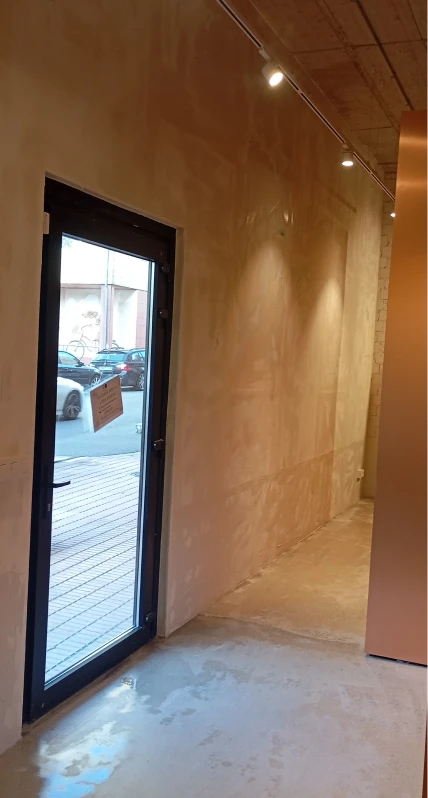 La imagen que has subido muestra una zona de una tienda o local comercial que aún está en proceso de finalización. El espacio tiene un suelo de cemento y las paredes están preparadas pero no pintadas, con una luz cálida iluminando la habitación desde el techo. A través de la puerta, se puede ver una calle con vehículos estacionados.
