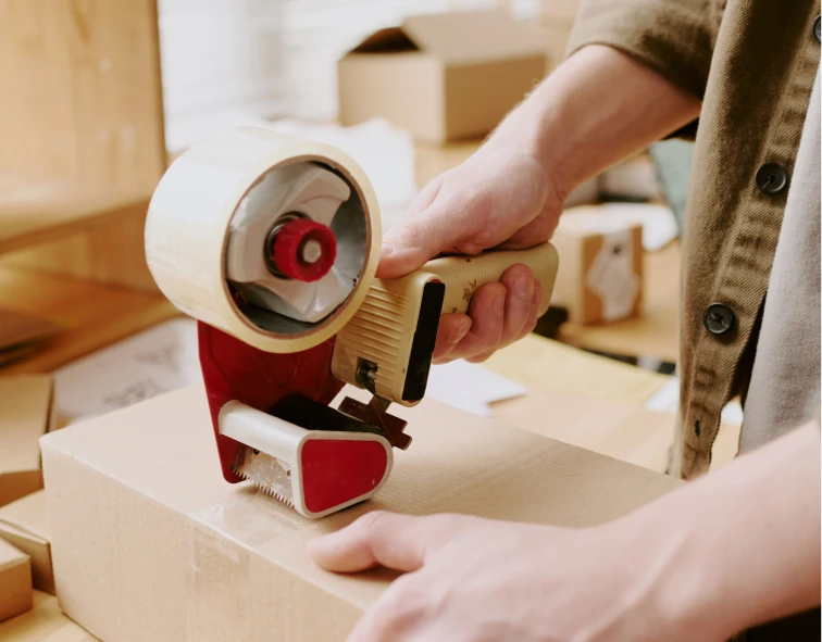 Mano usando una pistola de cinta adhesiva para sellar una caja de cartón, mostrando la precisión en el embalaje. El entorno de trabajo está ordenado, con más cajas en el fondo, lo que refleja un proceso de mudanza o preparación para el transporte.