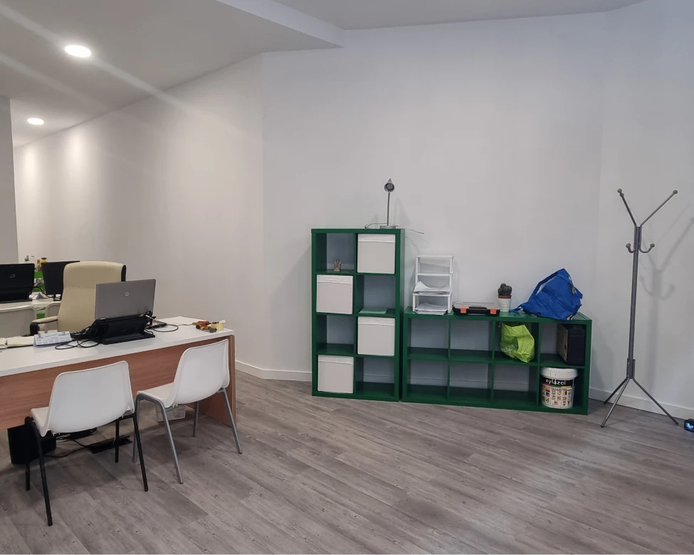 Oficina moderna y funcional, con un escritorio blanco y dos sillas minimalistas. A la derecha, estanterías verdes organizan el espacio de almacenamiento. El suelo de madera clara complementa la sencillez y eficiencia del diseño, creando un ambiente limpio y ordenado para un entorno de trabajo productivo.