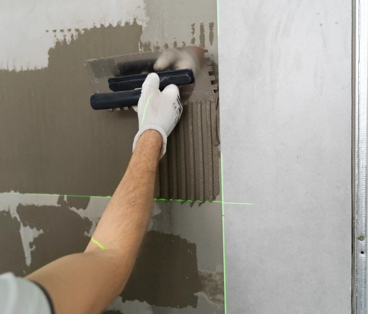 Obrero aplicando adhesivo en una pared utilizando una llana dentada, como parte de una renovación o instalación de azulejos. El uso de herramientas precisas y la atención a los detalles reflejan el profesionalismo en el proceso de reformas de interiores.
