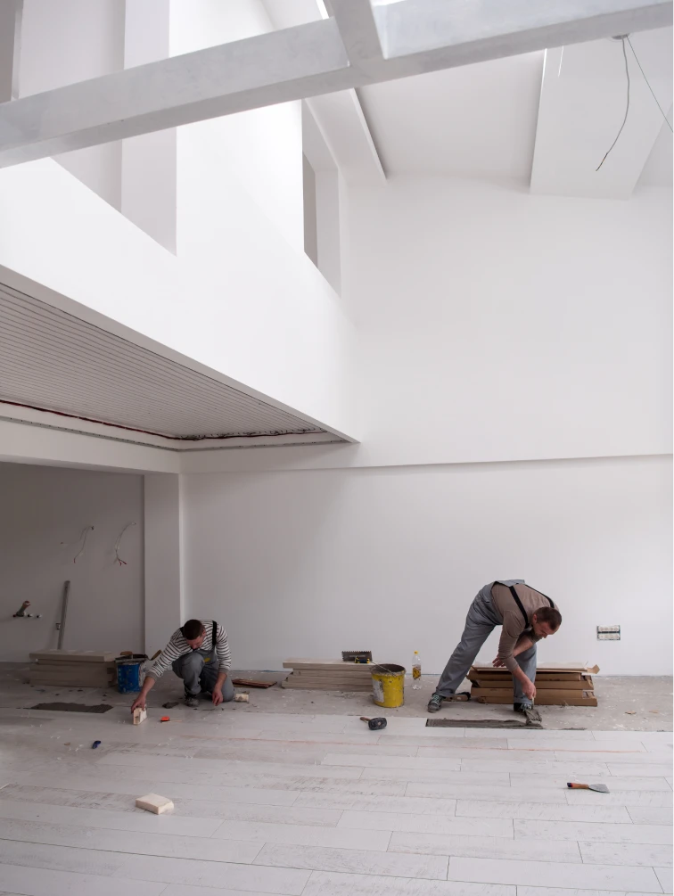 Obreros trabajando en una reforma de interiores, instalando suelo de madera en un espacio amplio y luminoso. La escena muestra a los trabajadores concentrados en su labor, utilizando herramientas profesionales para garantizar la precisión del trabajo en curso. El ambiente refleja un proceso de renovación en un espacio moderno con techos altos y paredes blancas.