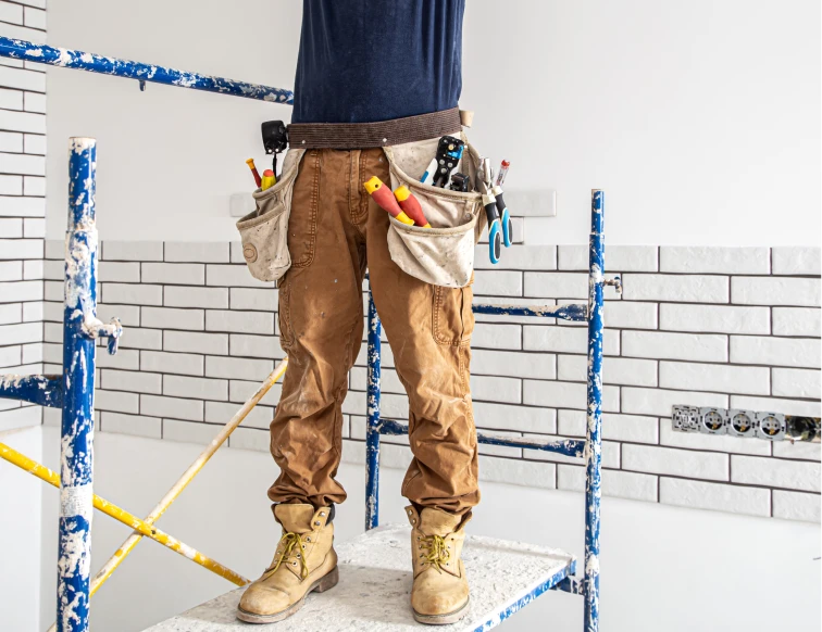 Trabajador de la construcción usando un cinturón de herramientas mientras está de pie sobre un andamio, rodeado de ladrillos blancos recién instalados en la pared. El cinturón de herramientas está lleno de diferentes utensilios de trabajo, lo que indica su preparación para continuar con la tarea de construcción o reforma.